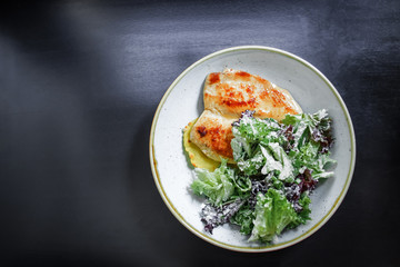 The main dish. Grilled chicken breast with a crust on a plate with mashed potatoes and green salad on a black background. Top view.