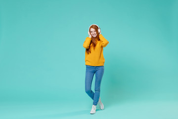 Pretty young redhead girl in yellow knitted sweater posing isolated on blue turquoise background studio portrait. People emotions lifestyle concept. Mock up copy space. Listen music with headphones.