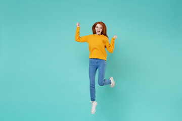 Happy young redhead woman girl in yellow knitted sweater posing isolated on blue turquoise background studio portrait. People lifestyle concept. Mock up copy space. Jumping doing winner gesture.