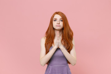 Pleading young redhead woman girl in plaid dress posing isolated on pastel pink background studio portrait. People sincere emotions lifestyle concept. Mock up copy space. Hold hands folded in preyer.
