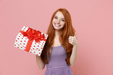 Smiling redhead woman girl in plaid dress isolated on pastel pink background. Valentine's Day Women's Day, birthday, holiday concept. Hold white red present box with gift ribbon bow, showing thumb up.