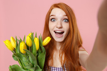 Close up of shocked redhead woman girl in plaid dress isolated on pastel pink background. People lifestyle concept. Mock up copy space. Hold bouquet of yellow tulips doing selfie shot on mobile phone.