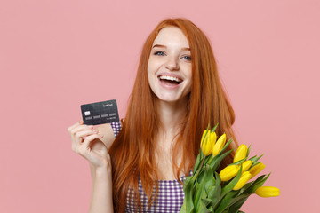Laughing young redhead woman girl in plaid dress posing isolated on pastel pink wall background in studio. People lifestyle concept. Mock up copy space. Hold bouquet of yellow tulips credit bank card.