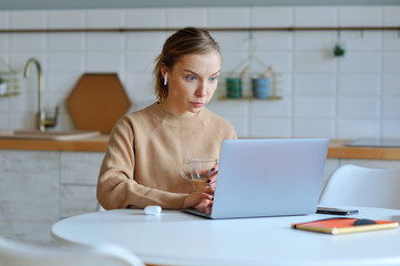 Lovely blonde woman working with her laptop while sitting in the kitchen in her apartment