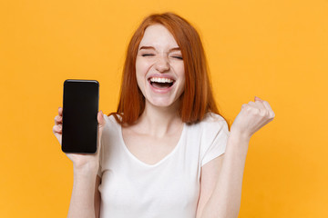 Joyful young redhead woman girl in white t-shirt posing isolated on yellow background. People lifestyle concept. Mock up copy space. Hold mobile phone with blank empty screen, doing winner gesture.