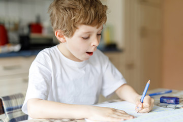 Hard-working happy school kid boy making homework during quarantine time from corona pandemic disease. Healthy child writing with pen, staying at home. Homeschooling concept