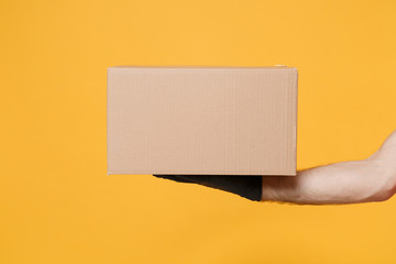 Close up cropped male hold in hand black glove brown clear blank paper empty cardboard box isolated on yellow background Packaging template mock up Delivery service concept Copy space Advertising area