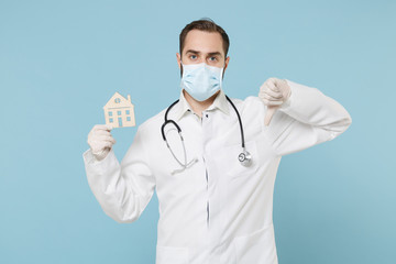 Male doctor man in medical gown face mask gloves isolated on blue background. Epidemic pandemic coronavirus 2019-ncov sars covid-19 flu virus. Hold house recommending stay home showing thumb down.