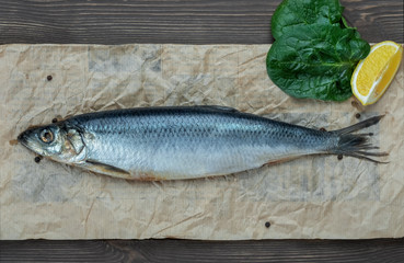 Healthy seafood, one whole salted atlantic herring on parchment paper, top view. Counter with fish in the market, wholesome diet food, next to spices, limon and lettuce leaf.