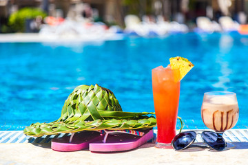 Summer vacation concept. Green hat from palm leaves, pink flip flops, sunglasses and cocktails near swimming pool tropical resort.