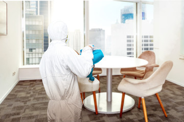 Man in a white protective suit spraying disinfectant in the office room - obrazy, fototapety, plakaty