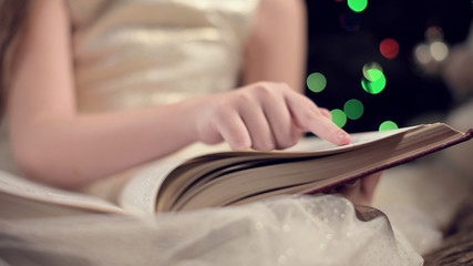 Close-up soft focus the hand of a little girl leads along the lines of a book while reading a New Year's fairy tale amid the lights of a Christmas tree. The concept of a fabulous mood
