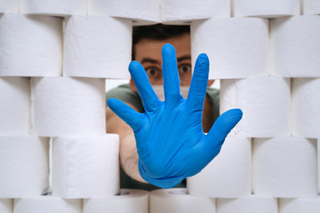 Closeup young caucasian man hidind behind the wall of tissue toilet paper during Coronavirus outbreak and showing stop gesture to Covid-19 spreading. Toilet paper background. Home isolation concept © Arsenii