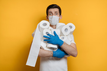 Shocked young man wearing protective medical face mask and gloves grabs many rolls of toilet paper, scared of Coronavirus outbreak. Covid-19 quarantine, social isolation, stay home concept. Copy space