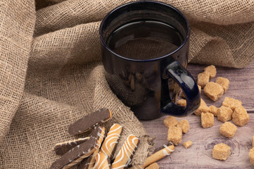 dark blue ceramic tea mug, cookie sticks with chocolate and white icing and pieces of brown cane sugar on a background of coarse-textured fabric. Close up.