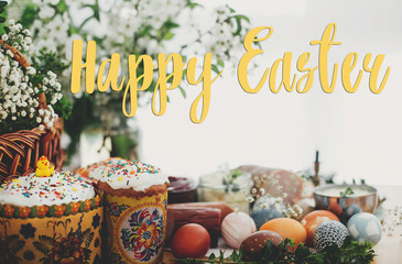 Happy Easter text. Easter greetings lettering. Easter modern eggs, easter bread, ham, beets, butter, cheese, green branches and flowers on rustic table. Greeting card