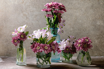 Group of glass bottles and vases with pink decorative flowers bouquets over grey texture background.