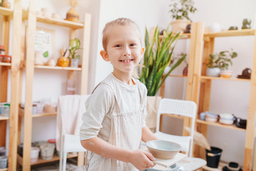 Cheerful boy in an apron with a clay plate in his hands, in pottery studio.