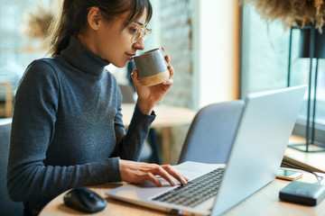 Young beautiful woman in eyeglasses enjoying coffee during work on portable, laptop, computer....