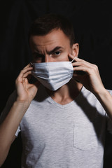 Apathetic man in medical mask from infection. Sad stressed male putting on fabric respirator from dangerous virus covid 19 while standing in dark studio on black background and looking at camera