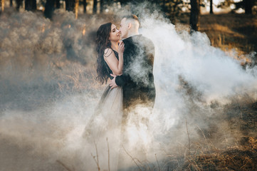 Fototapeta na wymiar A loving bridegroom in a black cardigan and a cute bride in an expensive dress are holding a pyrotechnic device with smoke in their hands and hugging. Wedding portrait of the newlyweds.