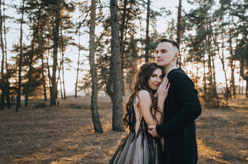 Stylish groom in a black cardigan and a beautiful bride in an expensive dress are hugging in the forest on the nature. Wedding portrait of smiling newlyweds. Photography, concept.