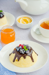 Cottage cheese casserole or pudding with chocolate sauce and berries on top on a white plate with a cup of tea on a light background. Healthy breakfast. Vertical orientation.