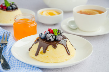Cottage cheese casserole or pudding with chocolate sauce and berries on top on a white plate with a cup of tea on a light background. Healthy breakfast. Horizontal orientation. Close up.