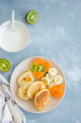 Pancakes on buttermilk or kefir with fresh fruit and yogurt on a plate on a light blue concrete background. A delicious and healthy breakfast. Vertical orientation. Top view, copy space.