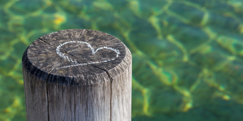 Naklejka na ściany i meble Starnberg, Bavaria / Germany - Mar 27, 2020: Chalk drawing of a heart on a rustic wooden pole / pile. In the background green colored water. With copy space. Card motif for love, marriage, valentine.