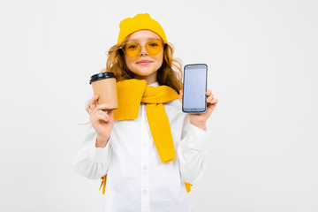girl in a winter hat and scarf with a glass of hot drink and a smartphone in her hands on a white background with copy space