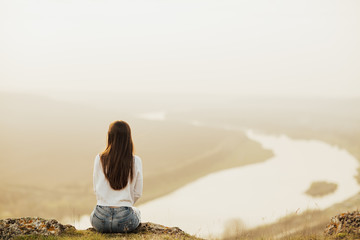 Back view of  woman sitting on top of the hill and looking at river in evening sunset. Young  traveler enjoying time in nature. Lifestyle travel, relaxation and freedom concept. Copy space. 