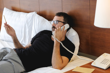Young businessman talking at the phone in hotel room ordering room service during self-isolation in quarantine - obrazy, fototapety, plakaty