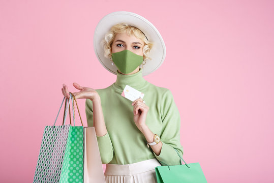 Spring online  shopping during quarantine conception: fashionable woman wearing protective mask posing with colorful paper bags and plastic bank card. Pink background. Copy, empty space for text