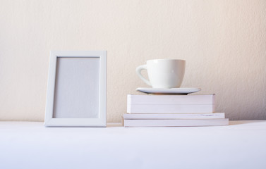 Fototapeta na wymiar White frame mock-up. Blank frame with white cup books by the white wall. Mock up frame concept. Minimalism concept. White concept