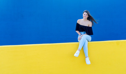 One young beautiful woman with long hair sitting on yellow wall while looking happy to camera in a sunny day