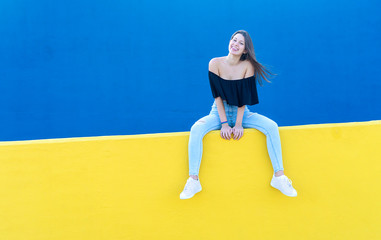 One young beautiful woman with long hair sitting on yellow wall while looking happy to camera in a sunny day
