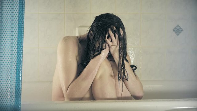 A depressed woman in the shower sits curled up on the ground and crying with her hands in her face as the water pours over her.