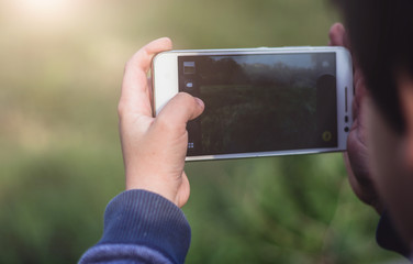 Kid holding a smartphone using both hands and taking a photo countryside landscape. Colorful sunny outdoors background, travel concept, close up.