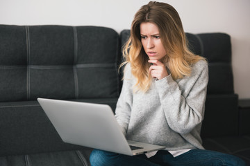 young blond beautiful woman sits on the sofa working from home on her laptop during quarantine looks thoughtful, cozy home atmosphere concept