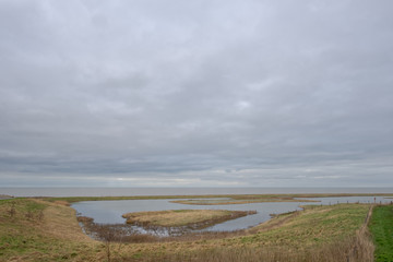 Large pool with water and small islands behind a Wadden Sea dike under a gray cloudy sky