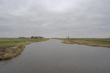 Wide ditch in a peat meadow landscape with a small windmill and a farm