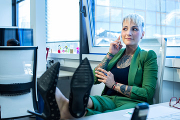 Segretaria con i capelli biondi , rasati ai lati , vestita con un completo verde e con diversi tatuaggi sul corpo, prla al cellulare seduta comodamente nella poltrona del suo ufficio