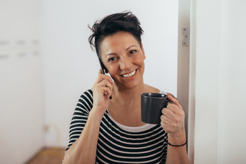 beautiful middle aged woman using mobile phone and drinking coffee in her home