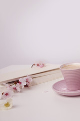 cup of cappuccino on a white table, blooming magnolia buds, a book in a white cover on a delicate pink background, a blank for the designer, an invitation form, a spring mood concept