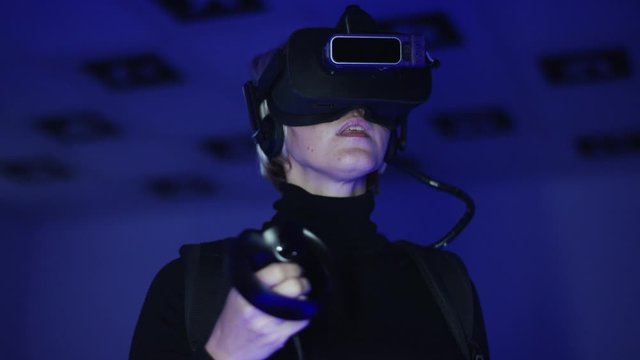Close up shot of an adult woman watching video in virtual realty headset with controller in her hand. Empty playroom full of trendy blickering red and blue neon lights. Tracking arc shot, 360 degree