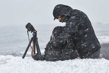 Photographer changing lens in challenging conditions in a snow storm using a beached iceberg as a...
