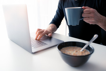 Middle-aged Caucasian man having breakfast while working from home, using his laptop, due to covid-19 coronavirus.
