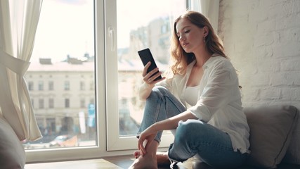 Attractive young woman sitting on a windowsill at home and texting on her phone communication female looking message cellphone cheerful smile use internet modern smartphone