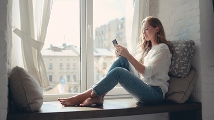 Pretty young woman sitting on a windowsill at home and texting on her phone communication female looking message cellphone cheerful smile use internet modern smartphone portrait